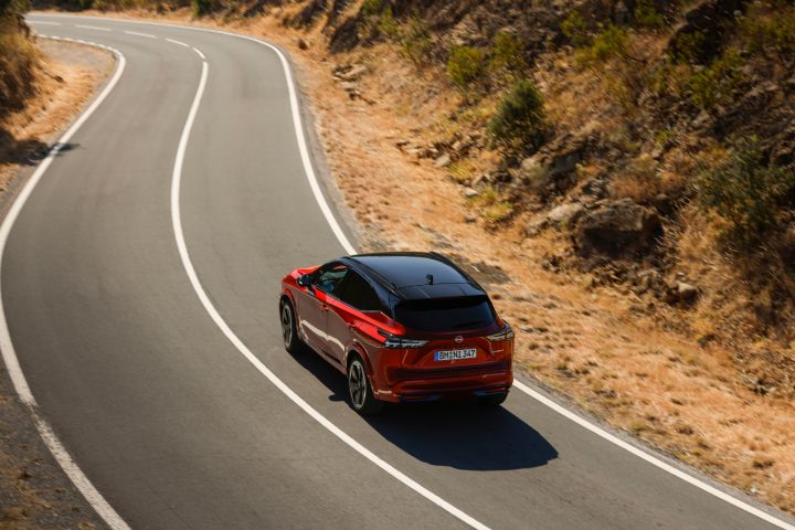 Nieuwe Nissan Qashqai in de kleur Fuji Sunset Red, van de achterzijde gezien, rijdend op een bochtige weg