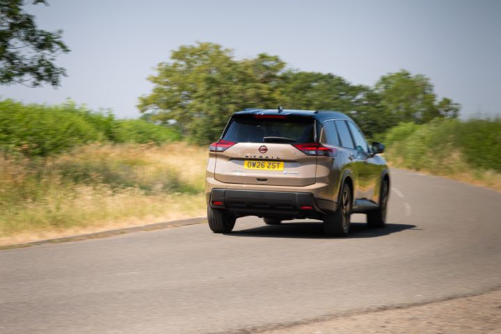Nissan X‑Trail driving along a country road, viewed from the rear three-quarter angle, with trees and grassland in the background.