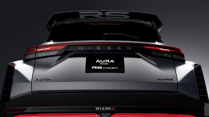 A close-up studio shot of the rear section of the Aura Nismo RS Concept, a silver and black two-tone sports car. The large rear wing features white lettering reading 'RS,' while the black plate at the center displays 'AURA NISMO RS CONCEPT' in white text. The taillights form sharp red lines across the rear, and a red accent line with the 'NISMO' logo is visible on the lower bumper. The background is a dark gray, emphasizing the car's detailed design