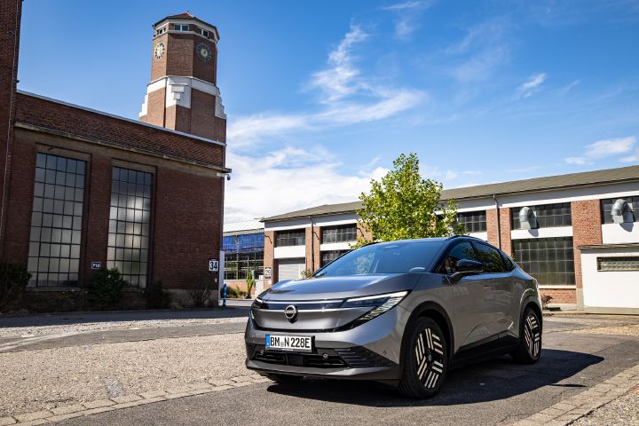 3/4 Frontansicht eines grauen Nissan Leaf, der in einem Innenhof zwischen Industrieanlagen mit Backsteinfassade und einem großen Turm steht
