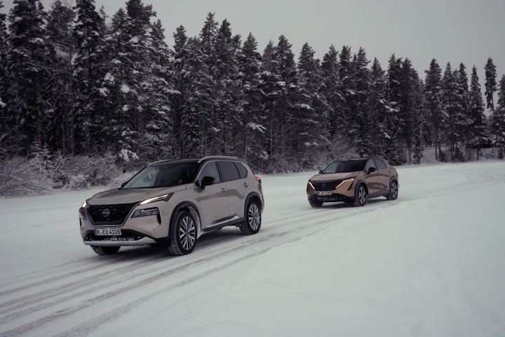 Silberner Nissan X‑Trail e‑Power e‑4ORCE und Elektroauto Nissan Ariya e‑4ORCE in Akatsuki Copper mit Dach und Außenspiegel in Pearl Black fahren hintereinander der auf schneebedeckter Straße mit verschneitem Wald im Hintergrund, 3/4 Front