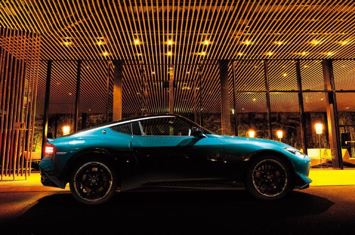 Unryu Green Fairlady Z parked in front of a modern illuminated building at night, photographed from the left-side profile. The vehicle faces toward the right side of the image, showcasing its long-hood, short-deck proportions, muscular rear fenders, and black alloy wheels. In the background, a glass-fronted structure with wooden slat architectural details is warmly lit, casting golden reflections across the vehicle and surrounding area. The rich Unryu Green body color contrasts with the ambient lighting, highlighting the refined styling and strong presence of the Fairlady Z.