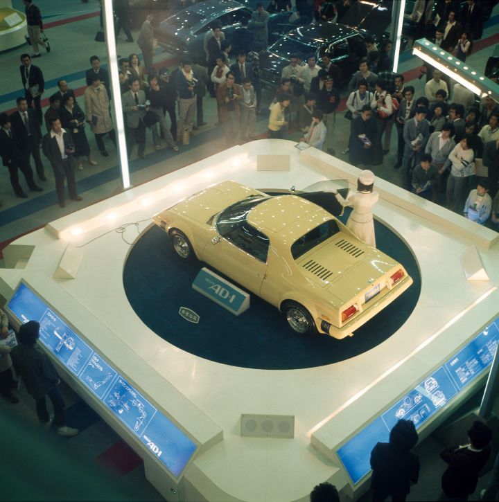 A vintage yellow Nissan car displayed at an auto show. a woman in white stands next to the open car door presenting.