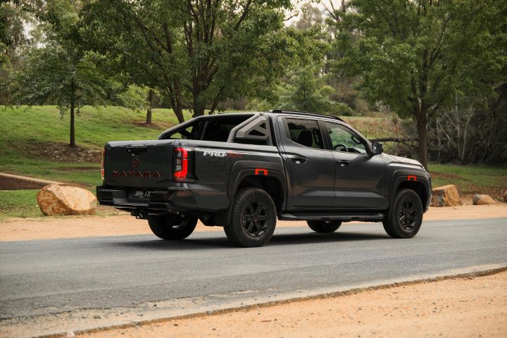 2026 Nissan Navara PRO-4X finished in Boulder Grey shown from the rear three-quarter angle with NAVARA-stamped tailgate, sports bar, tow hitch, black wheels and vertical tail-lights.