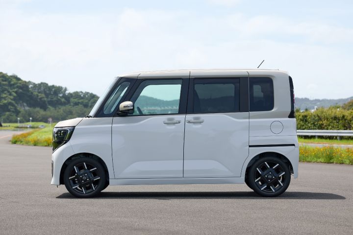 The new Nissan Roox kei car, in white with black wheels and tinted windows, is parked side-facing on a paved surface. The vehicle features a boxy design, and the background includes green trees and grass.