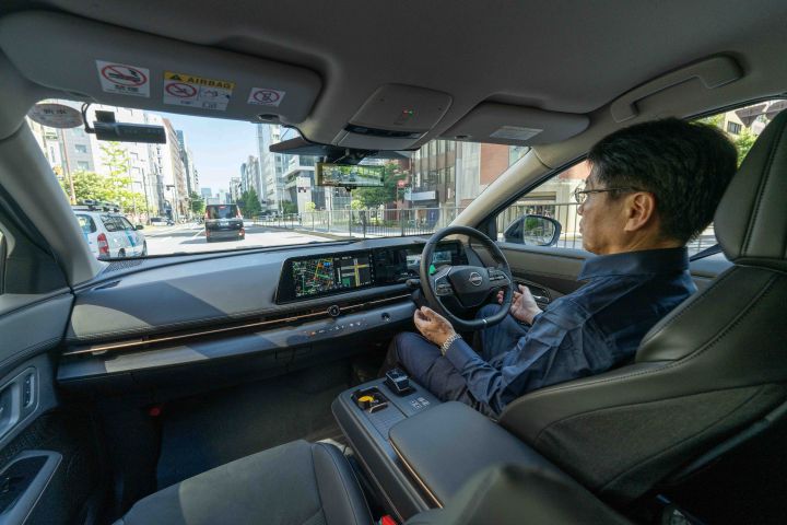 Innenansicht eines Nissan Ariya. Der Innenraum ist in Schwarz und Grau gehalten und verfügt über ein digitales Display auf dem Armaturenbrett. Eine Person sitzt auf dem rechten Fahrersitz und blickt nach vorne. Im Hintergrund ist eine Stadtlandschaft mit Straßen und Gebäuden zu sehen.