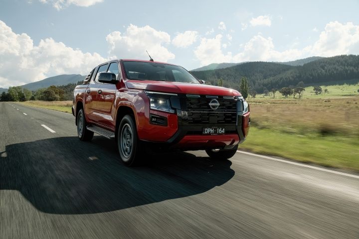 Three‑quarter front view of a 2026 Nissan Navara ST‑X Outback Red dual cab driving on a paved rural road framed by trees and dry roadside grass.