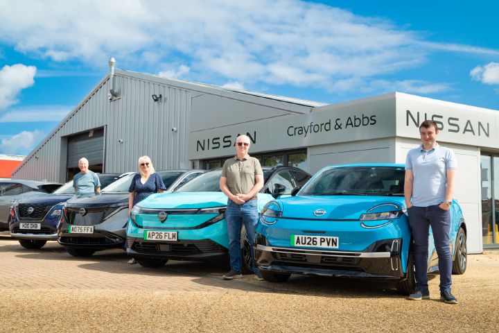 People standing beside a Nissan X‑Trail, Ariya, LEAF and MICRA outside a Nissan dealership