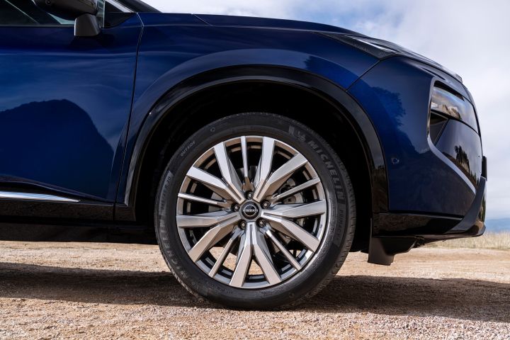 Neuer 2026 Nissan X‑Trail Tekna in der Farbe Universal Blue, Nahaufnahme des Vorderrads und Reifens auf Schotteruntergrund. Das Rad verfügt über eine silberne Mehrspeichenfelge aus Leichtmetall mit einem Nissan-Logo in der Mitte, und auf der Seitenwand ist die Reifenmarke „Michelin“ zu sehen.