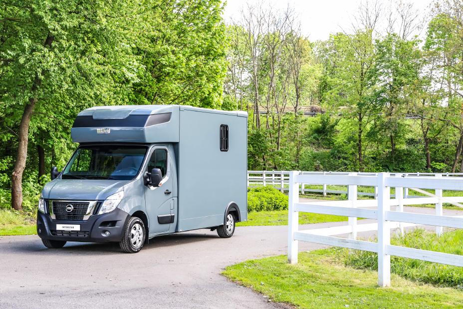 Grau-blauer Nissan Interstar Pferdetransporter von schräg vorne auf einem Weg zwischen zwei Pferdekoppeln mit weißem Gatter