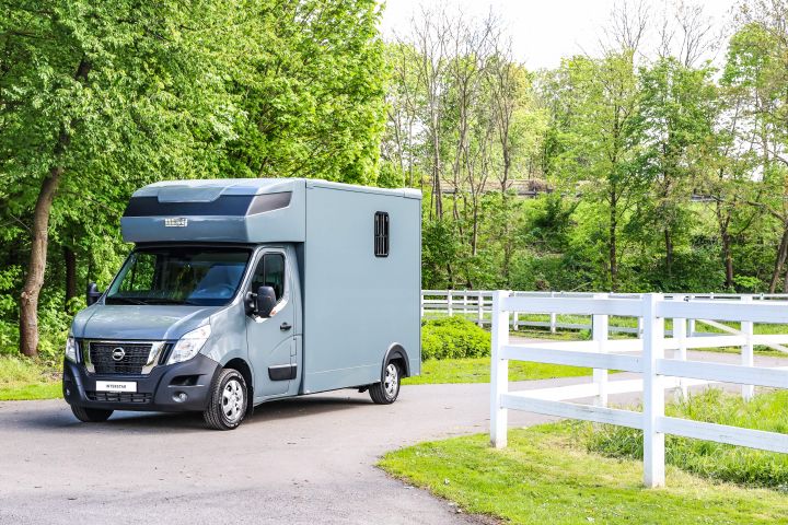 Grau-blauer Nissan Interstar Pferdetransporter von schräg vorne auf einem Weg zwischen zwei Pferdekoppeln mit weißem Gatter