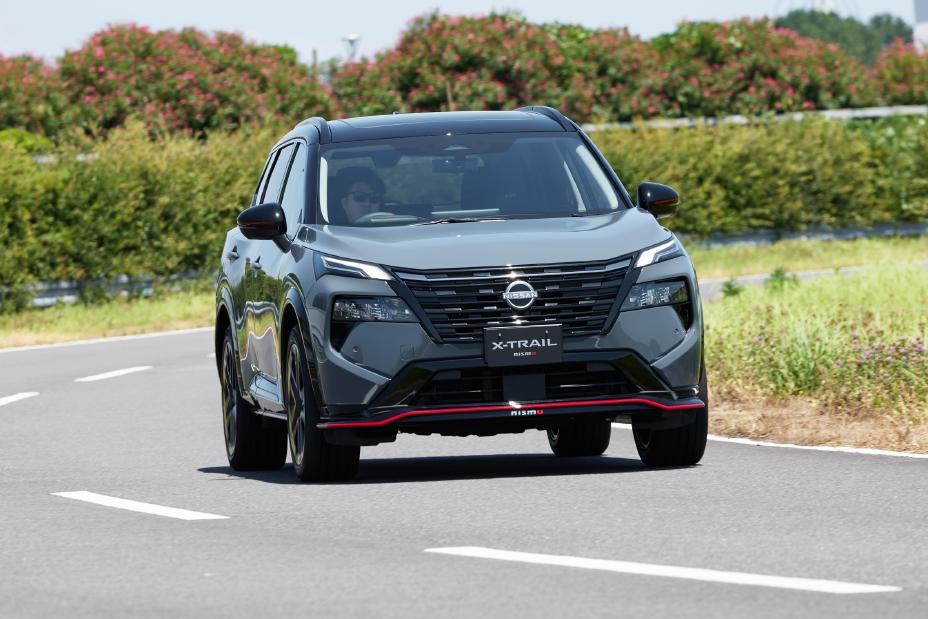 A grey Nissan X-Trail NISMO is driving on a road toward the camera. The road has white lane markings, and the background features trees and greenery.