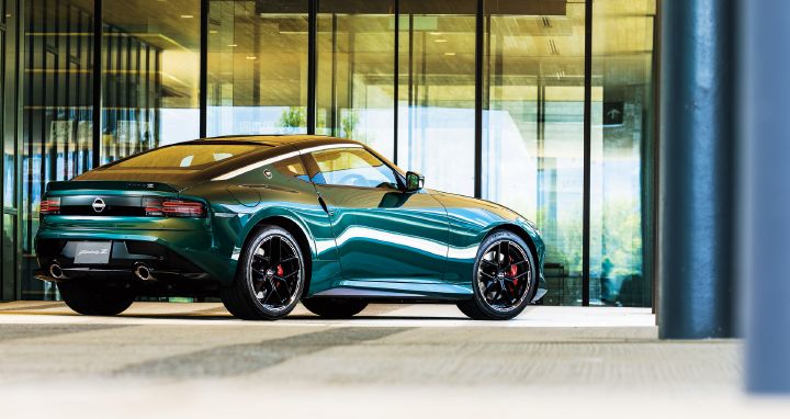 Unryu Green Fairlady Z parked in front of a modern glass-fronted building, photographed from a rear-left three-quarter angle. The vehicle faces toward the right side of the image, showcasing its distinctive rear combination lamps, muscular rear fenders, black alloy wheels, red brake calipers, and dual exhaust outlets. Large glass panels and dark structural columns form the background, while reflections from the building are mirrored across the body side. Bright natural light highlights the rich Unryu Green exterior finish and the flowing coupe silhouette, emphasizing the refined styling of the Fairlady Z.
