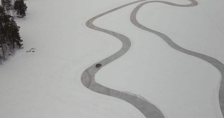 Silberner Nissan X‑Trail e‑Power e‑4ORcE fährt auf schneebedecktem Eissee mit verschneiten Bäumen und Holzhütten im Hintergrund, kleines Auto in weißer Landschaft, Drohnen-Perspektive