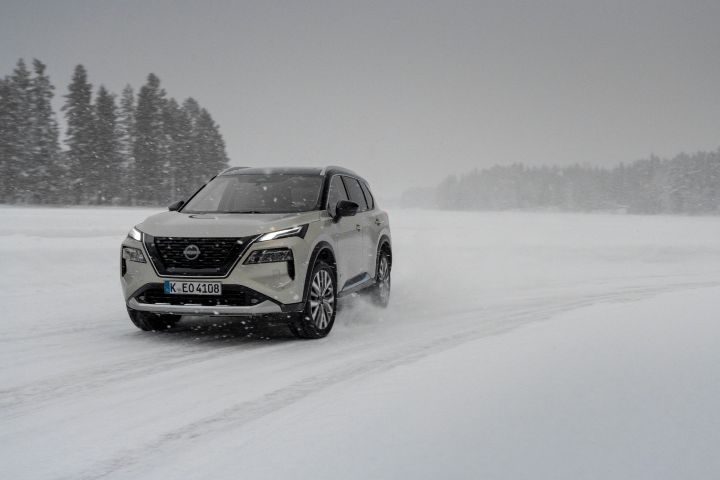 Silberner Nissan X‑Trail e‑Power e‑4ORcE driftet auf Eissee durch den Schnee mit verschneiten Bäumen im Hintergrund, 3/4 Front