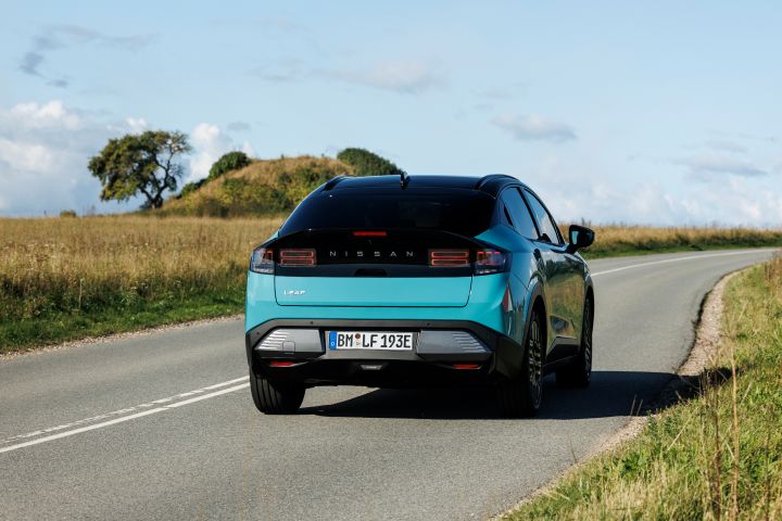 3/4 Heckaufnahme eines türkisen Nissan LEAF, der bei Sonnenschein und blauem Himmel über eine dänische Landstraße fährt