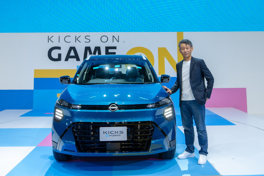 A blue car displayed on a brightly lit stage with geometric blue, white, and pink patterns. An executive stands beside the car, and the backdrop features large text that reads “GAME ON.”