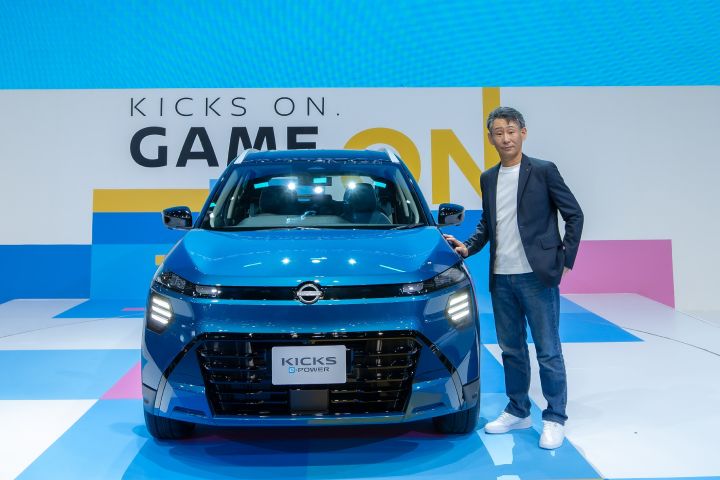 A blue car displayed on a brightly lit stage with geometric blue, white, and pink patterns. An executive stands beside the car, and the backdrop features large text that reads “GAME ON.”