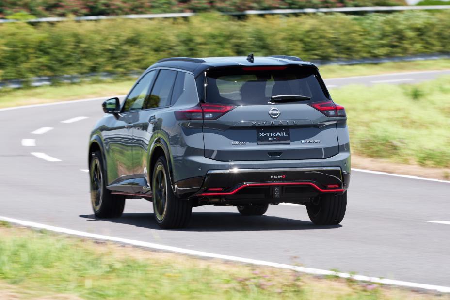 A grey Nissan X-Trail NISMO is driving on a curved road. The vehicle is captured from the rear, with greenery in the background.
