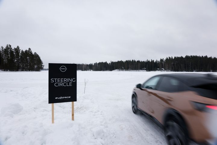 Schwarzes Schild mit weißer Schrift Nissan e‑4ORCE Steering Circle steht im Schnee, dynamische Seitenansicht des vorbeifahrenden Elektroautos Nissan Ariya e‑4ORCE in Akatsuki Copper mit Dach und Außenspiegel in Pearl Black