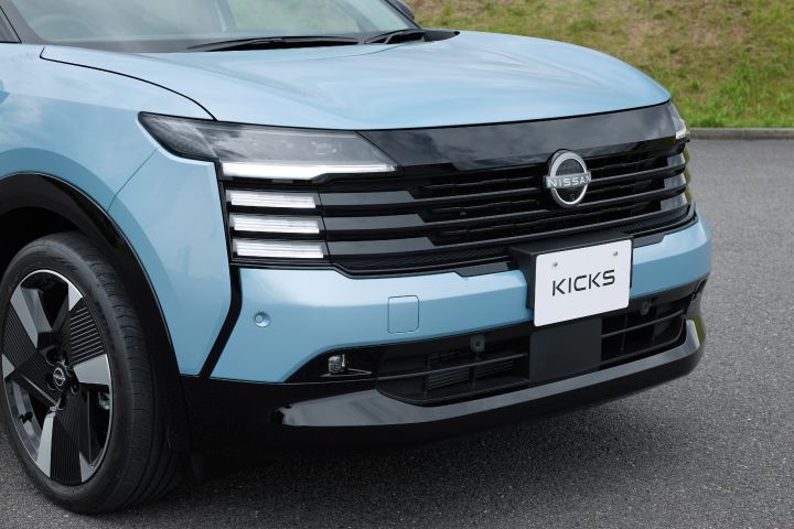 青みがかったキックスのフロント部分のクローズアップ。車両はやや右向きに配置され、フロントグリルとヘッドライトが強調されている。背景は舗装路と緑地。