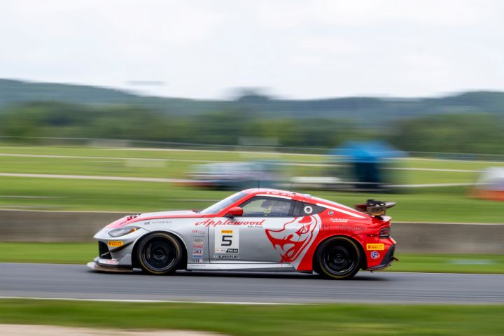 A silver and red Nissan Z GT4 race car racing on a track facing left with a blurry green background of trees and grass