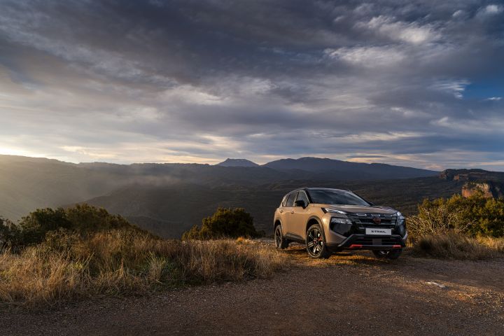 Der neue 2026 Nissan X‑Trail N-Trek in der Außenfarbe „Baja Storm“ steht auf einer unbefestigten Straße, umgeben von trockenem Gras und Sträuchern, mit einer bergigen Landschaft und einem dramatischen bewölkten Himmel im Hintergrund. Das Fahrzeug zeichnet sich durch elegante Designelemente mit schwarzen und magmaroten Akzenten aus, die seinen robusten und dennoch stilvollen Charakter unterstreichen, der sich für Offroad-Abenteuer eignet.