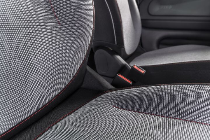 Foto, das eine Nahaufnahme des Fahrzeuginnenraums mit grauen Stoffsitzen mit Struktur, schwarzen Zierleisten und roten Ziernähten zeigt. Vordersitze, Rücksitze, Sicherheitsgurt-Schnallen und Türverkleidungen verdeutlichen die Polsterung und die Sicherheitsmerkmale des Fahrzeugs.