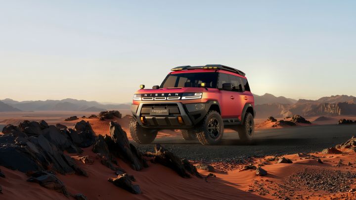 A red Terrano PHEV Concept driving at a front three-quarter angle across a rugged desert landscape. Rocks and sand appear in the foreground, with a sunset sky and distant mountains in the background.