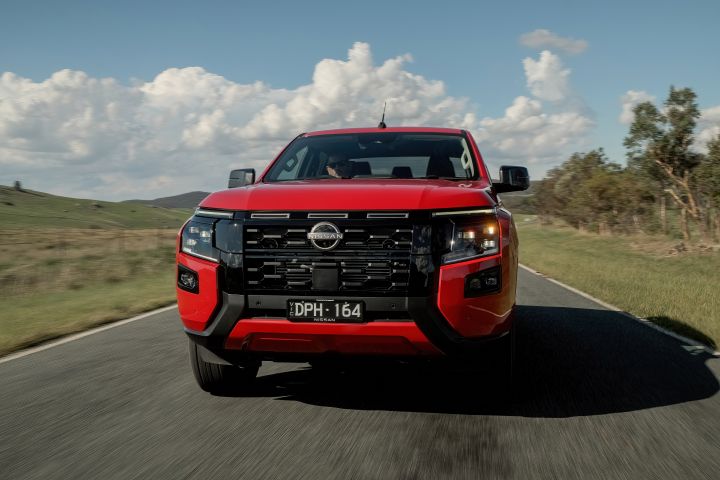 Front view of a red 2026 Nissan Navara ST‑X driving along a country road, with grassy hills and clouds in the background.