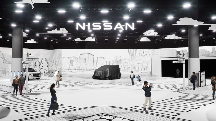 Ein Ausstellungsraum im Innenbereich verfügt über ein großes „NISSAN”-Logo an der Decke. In der Mitte ist ein mit einem schwarzen Tuch bedecktes Fahrzeug ausgestellt, umgeben von einer Kulisse aus schwarz-weißen Illustrationen, die ein städtisches Stadtbild darstellen. Der Boden ist mit Zebrastreifen- und Kreuzungsmustern gestaltet, und mehrere Personen gehen spazieren oder machen Fotos.
