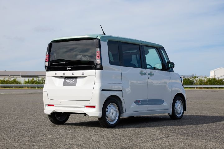 The new Nissan Roox kei car, in white, is parked rear-facing in a lot under a clear blue sky. The background includes paved ground and other vehicles.