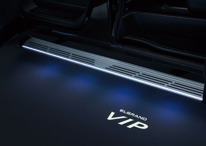 エルグランド VIPの助手席側スライドドア開口部下側をクローズアップで撮影した画像。画面上部にはブラックのボディと開いたスライドドアの下部が写っており、乗降口が開放された状態となっている。車両は側面から捉えられており、助手席とスライドドアに渡って、開口部にイルミネーション付き・シルバーのロングステップが水平に配置されている。
ロングステップの縁には白色のLEDラインが明るく発光しており、乗降時の足元を照らしている。画面下部の濃紺からブラックへと変化する床面には、白色の光で「ELGRAND VIP」の文字が投影されている。投影されたロゴは鮮明で、周囲には淡い青白い光が広がっている。
車内側にはブラックのフロアやドア開口部の内装が見え、落ち着いた高級感のある色調で統一されている。