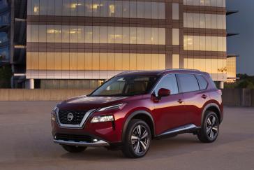 The front drivers side angle of a red Nissan Rogue with a large building in the background