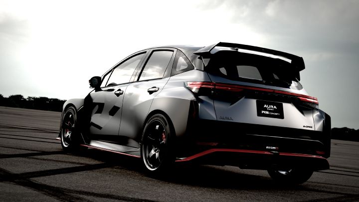 A silver and black two-tone sports car, the Aura Nismo RS Concept, parked on an outdoor skid pad in a rear-left angled view. The vehicle features a large rear wing, with a black plate displaying white text reading 'AURA NISMO RS CONCEPT' at the center of the rear, along with a red accent line and the NISMO logo on the lower bumper. The wheels are black with visible red brake calipers, and the side displays a light gray 'RS.' The background shows a cloudy sky and distant trees, with tire marks visible on the asphalt surface