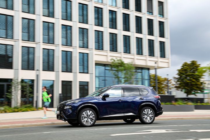 Seitliche Fahraufnahme eines dunkelblauen Nissan X‑Trail, hinter dem ein Mann mit hellgrünem Shirt vorbeijoggt