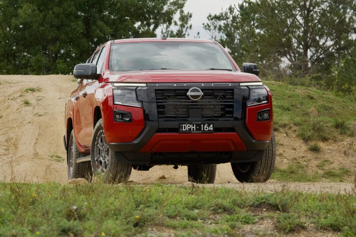 Low-angle front view of a 2026 Nissan Navara ST-X dual cab in Outback Red, positioned on a compacted dirt track between sandy banks with grass and trees in the background.