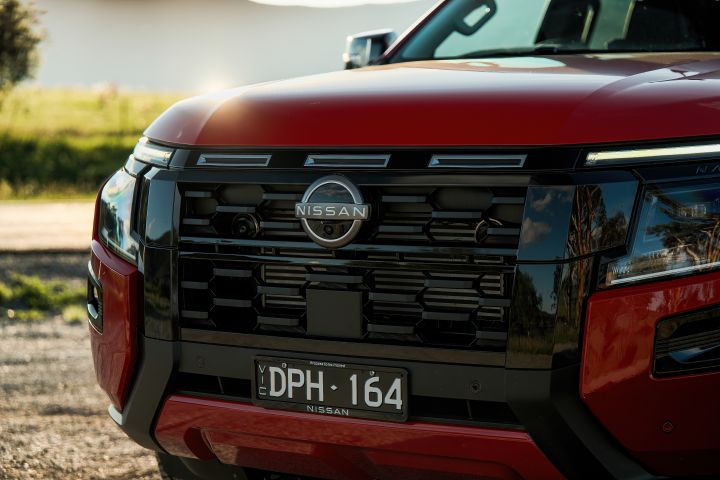 Front-end detail of a 2026 Nissan Navara ST-X in Outback Red, centred on the black grille, Nissan badge, front camera, number plate, and surrounding bumper elements.