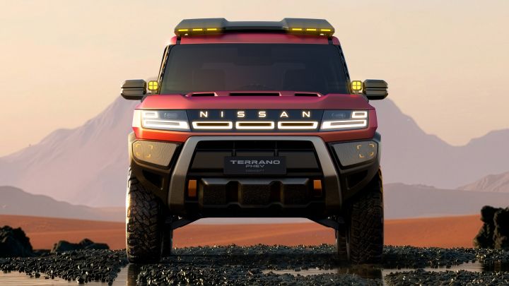 A red Terrano PHEV Concept parkng toward the viewer across a rugged desert landscape. A sunset sky and distant mountains fill the background, with rocks and sand visible on the ground.