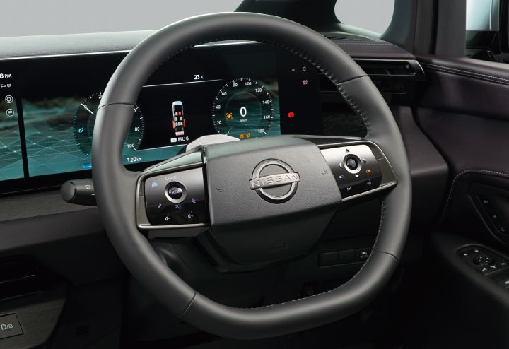 エルグランド AUTECH LINEの運転席まわりを、運転席側やや左後方からクローズアップで撮影した画像。画面中央にはブラックのステアリングホイールが配置され、中央に日産エンブレムが大きくあしらわれている。ステアリングには左右それぞれスイッチやタッチ式操作部が組み込まれ、先進的なデザインとなっている。
ステアリングの奥には横長の大型デジタルディスプレイが広がり、左側には車両情報画面、中央には速度計や運転支援表示を含むデジタルメーターが表示されている。メーターパネルには車両のドア開閉状態を示すイラストや速度表示「0 km/h」などが確認できる。
ダッシュボードはブラックを基調とし、右側にはエアコン吹き出し口やドアトリムの一部が見え、ドア内張りには濃いパープルがかった加飾とステッチが施されている。車両は右ハンドル仕様で、ステアリングは車内右側に配置されている。