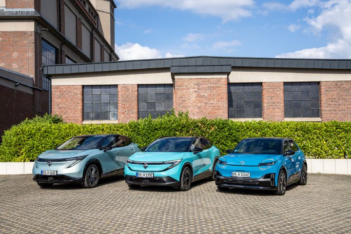 Vor einem Backsteingebäude und einer grünen Hecke stehen nebeneinander die drei Nissan EV-Modelle Nissan Ariya, Nissan Leaf und Nissan Micra, 3/4 Frontansicht aus leicht erhöhter Position