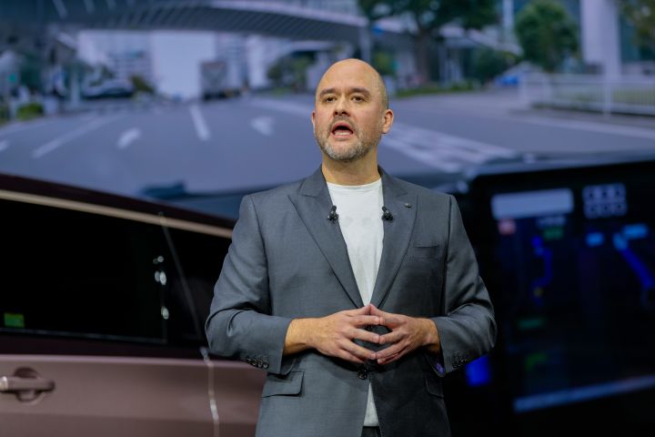 On the stage, CEO Ivan Espinosa, dressed in a suit, stands with both hands clasped in front while delivering a presentation. In the foreground, part of the all-new Elgrand is visible, and in the background, a large screen displays an urban road scene featuring lanes, elevated structures, and green landscaping.