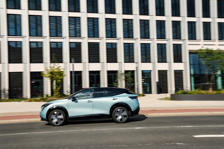 Vor einer hohen, hellen Fassade mit vielen Fenstern fährt seitlich ein hellblauer Nissan Ariya über eine sonnenbeschienene Straße