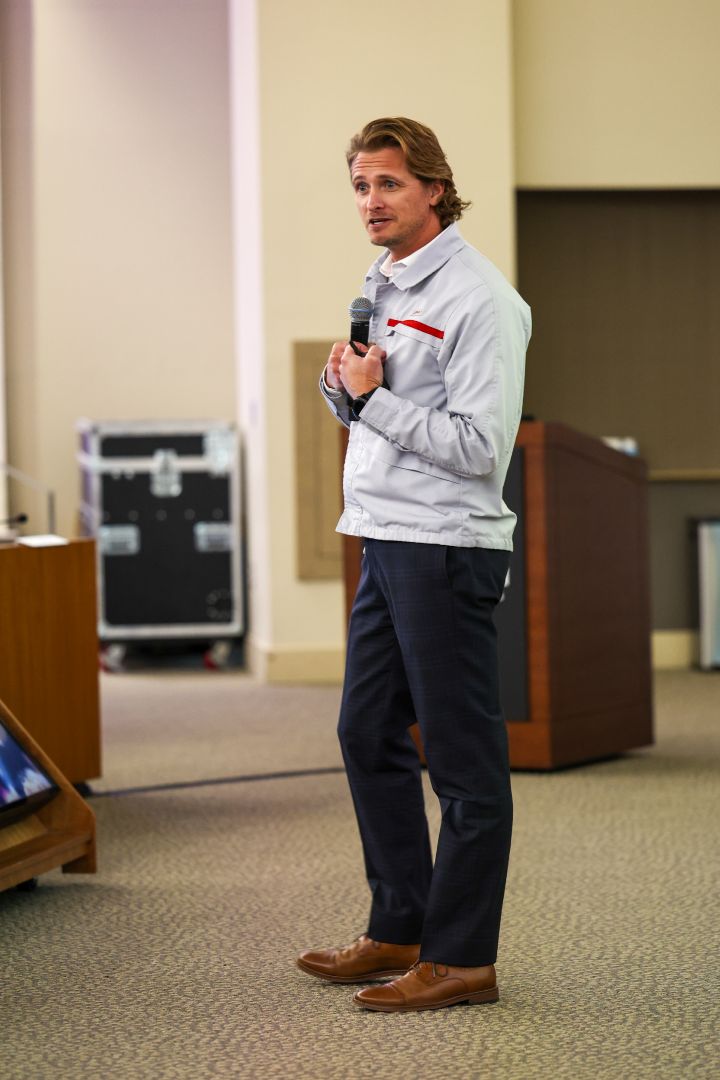A man in a white windbreaker speaking into microphone to a crowd.
