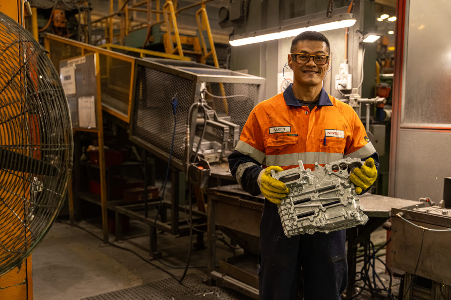Factory worker wearing high‑visibility orange protective clothing and yellow gloves holds a freshly cast metal automotive component inside an industrial manufacturing environment with machinery, safety railings, and overhead lighting.