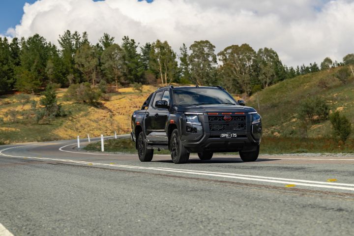 2026 Nissan Navara PRO-4X finished in Boulder Grey shown in a long-lens front three-quarter view approaching a broad curve with guide posts and ochre embankments.
