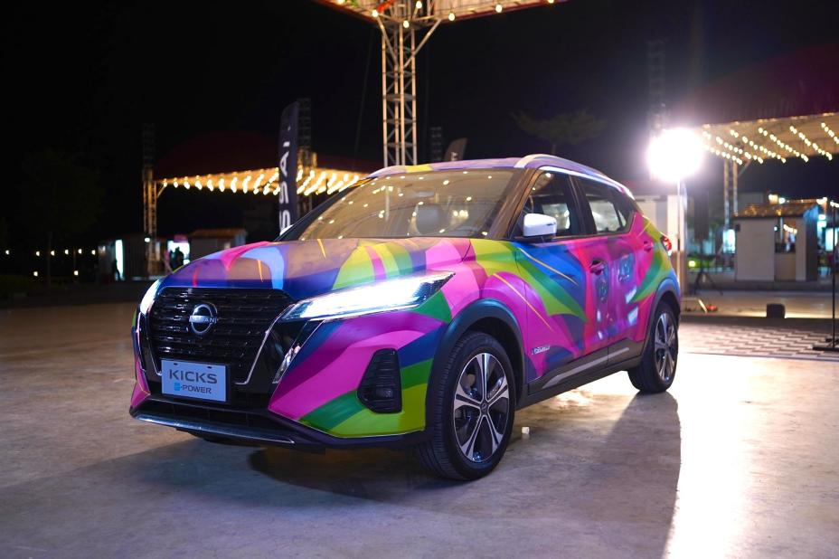 A Nissan Kicks e‑POWER displayed outdoors at night, featuring a vibrant and colorful graffiti-style wrap with bright pink, green, yellow, and blue designs under event lighting.