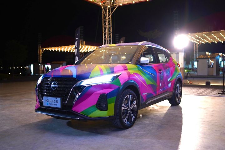 A Nissan Kicks e‑POWER displayed outdoors at night, featuring a vibrant and colorful graffiti-style wrap with bright pink, green, yellow, and blue designs under event lighting.