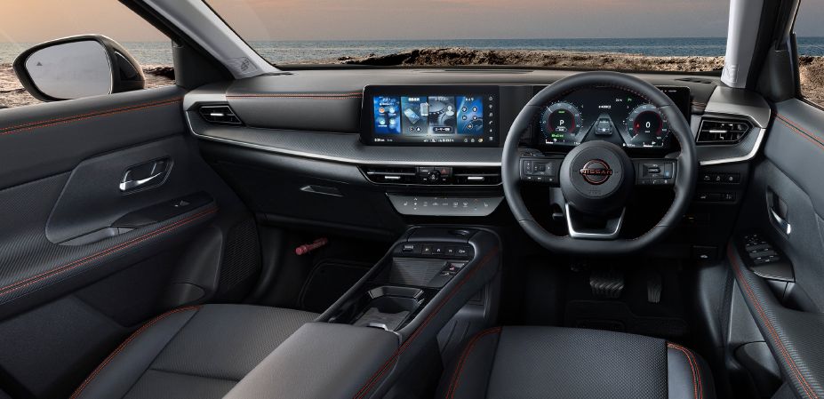 Interior view of the kicks rock creek from the driver's side. The black-themed cabin features a steering wheel on the right, a full digital meter display directly ahead, and a wide center display positioned to the left. Lava red stitching is applied to the dashboard, door trims, and seats, creating the exclusive outdoor-inspired character of the rock creek grade. Beyond the windshield, a rocky shoreline and the horizon are visible beneath a sky shaded in soft orange and gray tones.