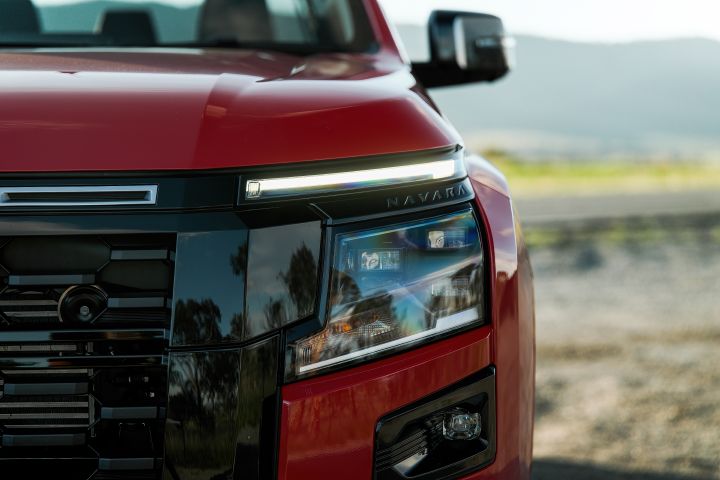 Close-up of the front headlamp and upper grille trim on a 2026 Nissan Navara ST-X in Outback Red, showing the illuminated daytime running light, NAVARA script, and black gloss fascia.
